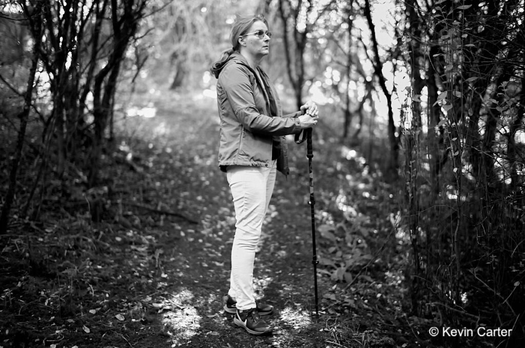 A photo of a woman holding a hiking pole to steady herself on rough ground. She is under the canopy of trees.