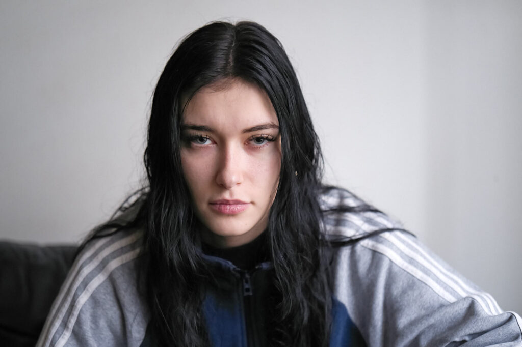 A young woman with dyed black hair sitting on a sofa in dreadfully low light.