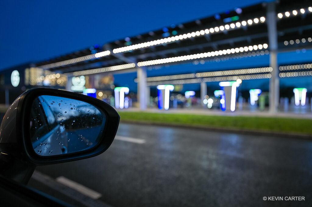 An electric charging forecourt with early evening skies and a rear view mirror